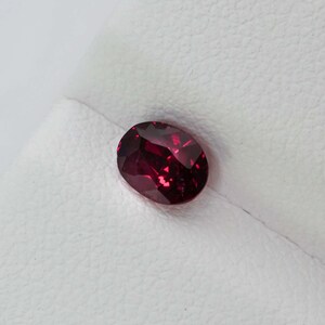 May include: A faceted, oval-cut, deep red gemstone. The gem reflects light, displaying its rich colour. The stone is set against a white background, highlighting its vibrant hue. This gemstone is suitable for jewellery making.