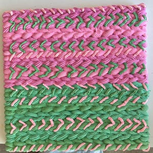 May include: A square, hand-woven textile in shades of pink and green. The design features rows of textured, braided patterns, with alternating colors creating a visually appealing, geometric effect. The piece is likely a decorative item or a small rug.