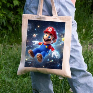 May include: A beige tote bag featuring a vibrant illustration of Mario in a starry space scene. Mario is depicted in his signature red hat, blue overalls, and white gloves, with a joyful expression. The background is filled with stars and shooting stars.