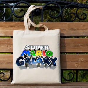 May include: A beige canvas tote bag featuring the words "SUPER MARIO GALAXY" in a colorful, 3D-style design. The bag has two handles and is sitting on a wooden bench.