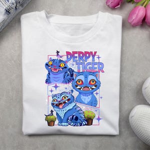 May include: White t-shirt featuring a colorful graphic of three blue cartoon tigers with the text "DERPY TIGER" in pink and blue. The tigers have exaggerated features and are surrounded by stars and small potted plants.