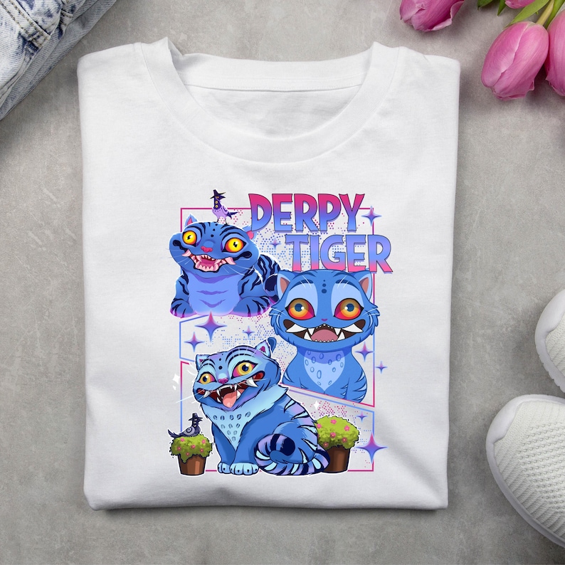 May include: White t-shirt featuring a colorful graphic of three blue cartoon tigers with the text "DERPY TIGER" in pink and blue. The tigers have exaggerated features and are surrounded by stars and small potted plants.