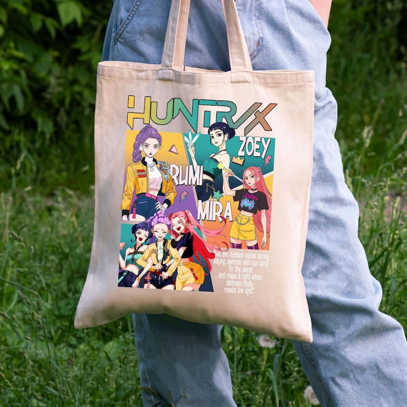 May include: Beige tote bag featuring a colorful anime-style graphic with the text "HUNTRAX" and the names "ZOEY", "RUMI", and "MIRA". The graphic includes illustrations of several anime characters in various poses and outfits. The bag has two handles.