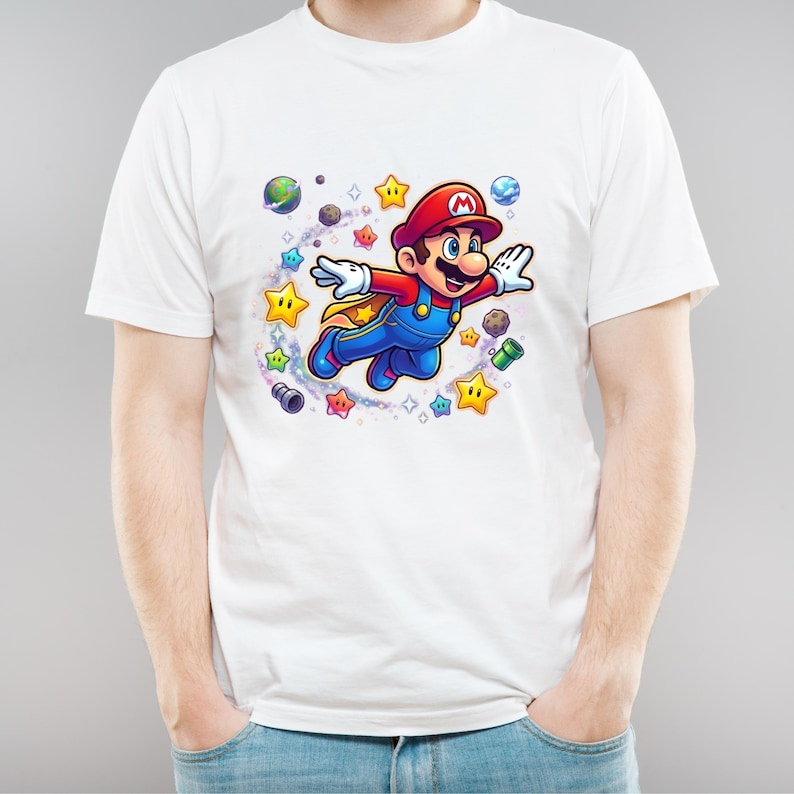 May include: White t-shirt featuring a colorful illustration of a flying cartoon character with a red hat, blue overalls, and a yellow cape. The design includes stars, planets, and other game-related elements.