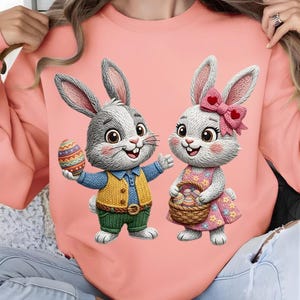 以下が含まれることがあります： 2匹のウサギの漫画イラストが描かれた、明るいピンク色のスウェットシャツ。1匹のウサギは装飾されたイースターエッグを持ち、もう1匹は卵のバスケットを持っています。ウサギはカラフルな衣装を着ており、片方のウサギの耳にはリボンが付いています。