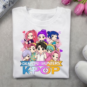 May include: White t-shirt featuring a colorful graphic of anime-style characters. The design includes the text "DEMON HUNTERS" above "KPOP" in a gradient color scheme. The characters have various hair colors and expressions.
