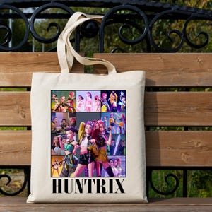 May include: A cream-colored tote bag featuring a colorful collage of images and the word "HUNTRIX" in bold, black letters. The collage includes images of people in various outfits and poses. The bag has a simple design with long handles.