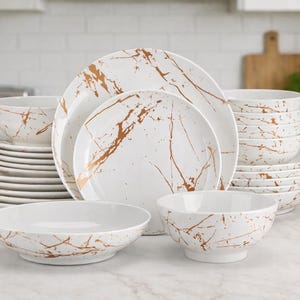 May include: A full white dinner service with a gold marble effect. The set includes plates and bowls of various sizes, neatly stacked. The dishes are arranged on a white countertop, ideal for a modern kitchen.