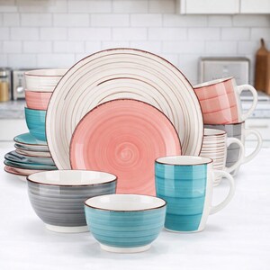 May include: A complete dinnerware set featuring plates, bowls, and mugs in various sizes. The set includes plates with a white and brown spiral design, pink and teal plates, and mugs with matching colors. The bowls are gray and teal with a white interior.