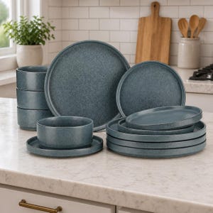 May include: A blue-grey speckled dinner set, including plates, bowls, and cups. The plates are stacked, and the cups are arranged in a neat stack. The set is displayed on a kitchen countertop, showcasing a modern design.