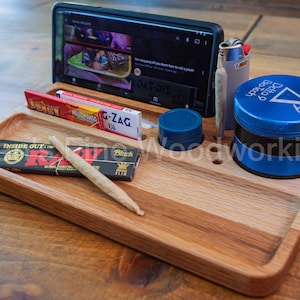 May include: A wooden rolling tray with a built-in phone stand, holding a smartphone displaying a video. The tray also holds rolling papers, a pre-rolled joint, a lighter, a blue grinder, and a blue cap.