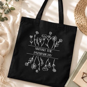Op de afbeelding: Zwarte draagtas met wit bloemmotief en de tekst "SMASH THE PATRIARCHY". De tas heeft twee zwarte handvatten en is gemaakt van een stevige stof. De tas ligt op een wit oppervlak met een kopje koffie en een boek.