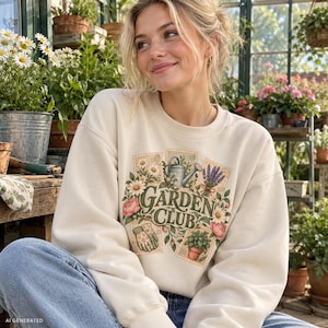 Könnte beinhalten: Ein cremefarbenes Sweatshirt mit Rundhalsausschnitt und einem Grafikdruck mit dem Text GARDEN CLUB. Das Design zeigt Illustrationen einer Gießkanne, Lavendel, Gänseblümchen, rosa Pfingstrosen, Gartenhandschuhen und einer Topfpflanze.