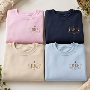 Könnte beinhalten: Vier Rundhals-Sweatshirts in Hellrosa, Marineblau, Beige und Hellblau. Jedes hat eine kleine Stickerei auf der linken Brust mit dem Text "BEE HAPPY", umgeben von Blumenranken, einer kleinen Biene und einem winzigen Herzsymbol.