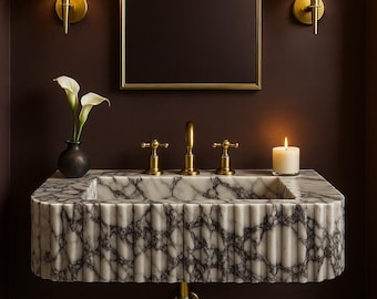 May include: A marble bathroom sink with a fluted design, gold fixtures, and a matching arched mirror. Two sconces with pleated shades flank the mirror. A small vase with white flowers and a lit candle add to the decor.