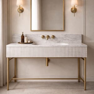 Puede incluir: Un tocador de baño moderno con un mueble blanco con ranuras verticales y una encimera de mármol. El tocador tiene un marco de metal dorado y un espejo arqueado a juego. Dos apliques flanquean el espejo y una bandeja con botellas se encuentra sobre la encimera.