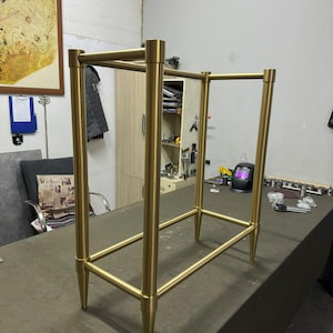 May include: A gold-coloured metal frame with a rectangular shape, featuring four vertical posts and horizontal bars. The frame has a minimalist design with tapered legs, suggesting a modern furniture piece. The structure appears to be sturdy and well-crafted.