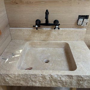 Puede incluir: Un lavabo de piedra natural beige con un borde frontal de acabado rugoso y una cubeta lisa, combinado con un grifo negro mate montado en la pared que presenta dos manijas en cruz con tapas centrales blancas, sobre un panel de madera clara.