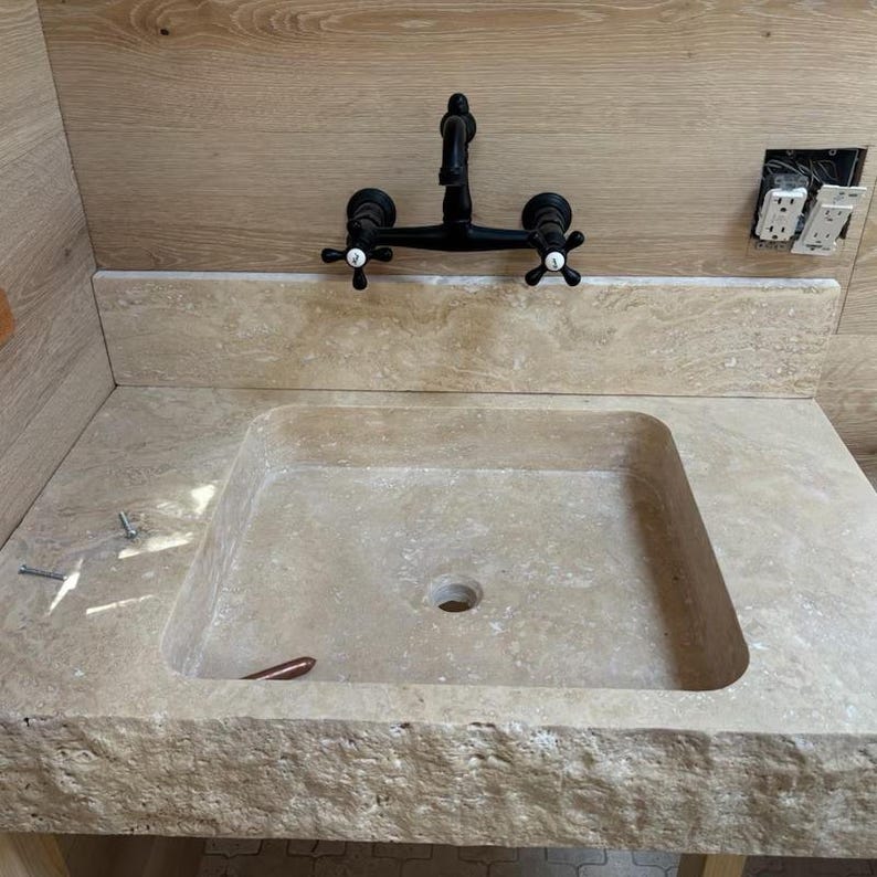 Puede incluir: Un lavabo de piedra natural beige con un borde frontal de acabado rugoso y una cubeta lisa, combinado con un grifo negro mate montado en la pared que presenta dos manijas en cruz con tapas centrales blancas, sobre un panel de madera clara.