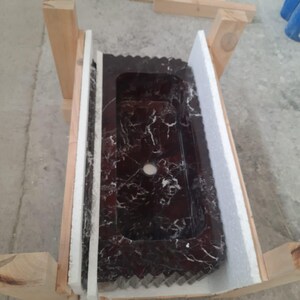 May include: A rectangular, dark red marble sink with white veining, set within a wooden frame and protected by white foam. The sink has a single centered drain hole and a decorative edge.