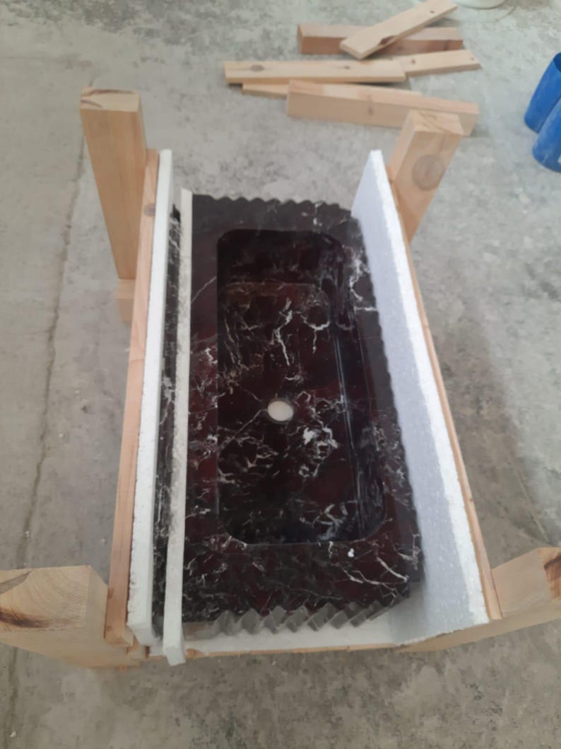 May include: A rectangular, dark red marble sink with white veining, set within a wooden frame and protected by white foam. The sink has a single centered drain hole and a decorative edge.