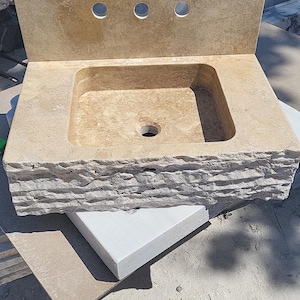 Puede incluir: Un lavabo de ba&ntilde;o de piedra beige con una cubeta rectangular, un salpicadero plano con tres orificios circulares para grifos y un borde frontal rugoso y cincelado. La cubeta tiene un orificio de drenaje central y es de piedra natural.