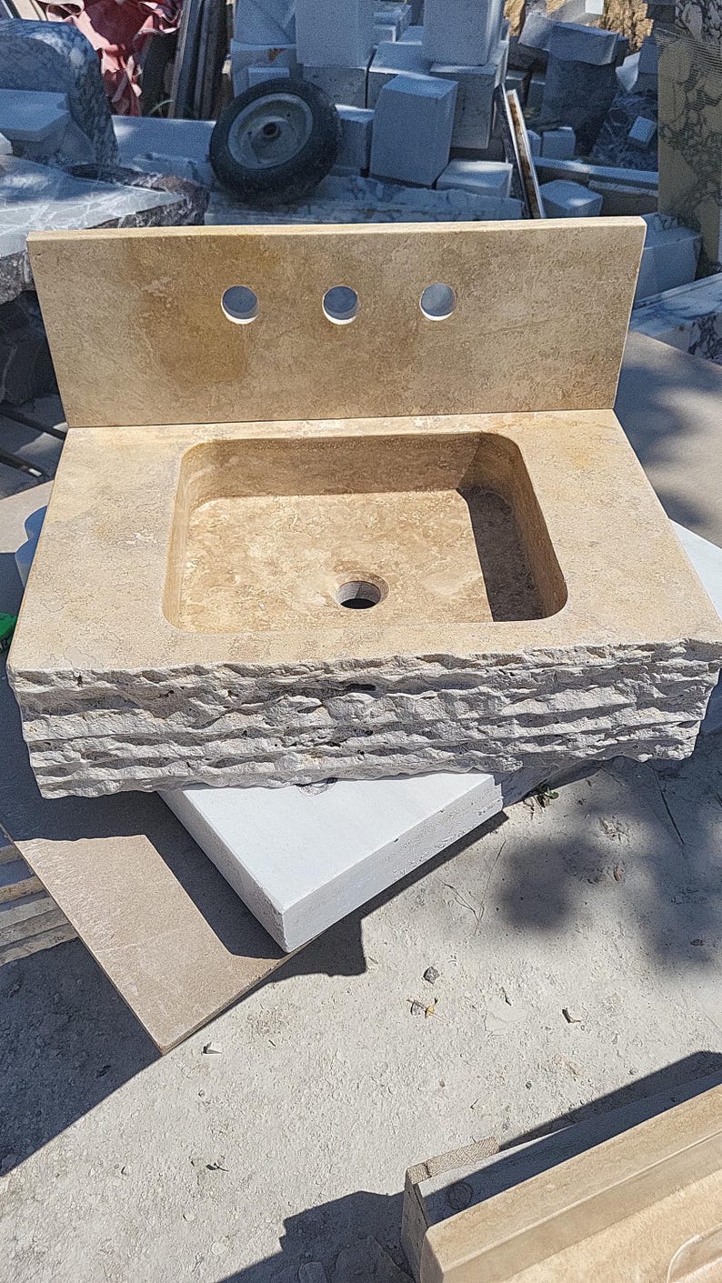 Puede incluir: Un lavabo de ba&ntilde;o de piedra beige con una cubeta rectangular, un salpicadero plano con tres orificios circulares para grifos y un borde frontal rugoso y cincelado. La cubeta tiene un orificio de drenaje central y es de piedra natural.
