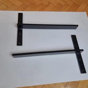 May include: Two black metal shelf brackets. Each bracket has a vertical support with pre-drilled holes and a horizontal arm. The brackets are designed for wall mounting and are suitable for supporting shelves.