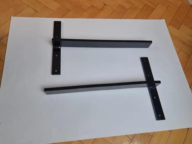 May include: Two black metal shelf brackets. Each bracket has a vertical support with pre-drilled holes and a horizontal arm. The brackets are designed for wall mounting and are suitable for supporting shelves.