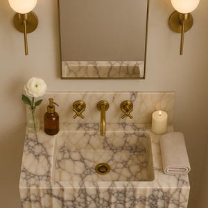 May include: A marble sink with gold fixtures, a mirror, and two wall sconces. The sink has a rectangular basin and a gold drain. A small candle, a rolled towel, and a soap dispenser are on the countertop. A white flower in a vase is also present.