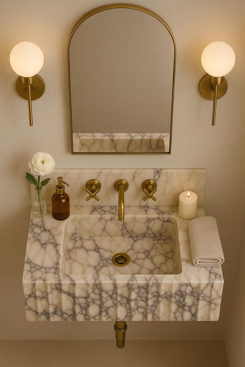 May include: A marble sink with gold fixtures, a mirror, and two wall sconces. The sink has a rectangular basin and a gold drain. A small candle, a rolled towel, and a soap dispenser are on the countertop. A white flower in a vase is also present.