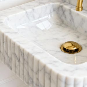 Puede incluir: Un lavabo de m&aacute;rmol blanco con un dise&ntilde;o acanalado en la parte delantera y los lados. El lavabo tiene una cubeta rectangular y una tapa de desag&uuml;e dorada. Un grifo dorado es visible en la esquina superior derecha. Es un accesorio de ba&ntilde;o.