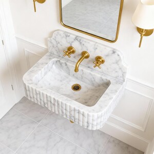 Puede incluir: Un lavabo de m&aacute;rmol blanco montado en la pared con un dise&ntilde;o acanalado. El lavabo tiene accesorios dorados, incluyendo un grifo y dos v&aacute;lvulas de manija en cruz. Un espejo con marco dorado y apliques con pantallas blancas est&aacute;n montados encima del lavabo. El suelo est&aacute; embaldosado.