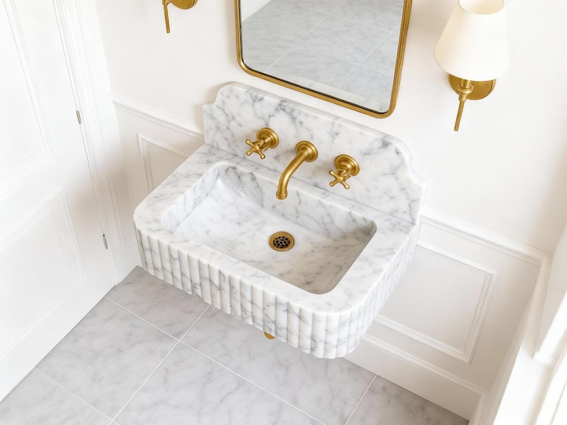 Puede incluir: Un lavabo de m&aacute;rmol blanco montado en la pared con un dise&ntilde;o acanalado. El lavabo tiene accesorios dorados, incluyendo un grifo y dos v&aacute;lvulas de manija en cruz. Un espejo con marco dorado y apliques con pantallas blancas est&aacute;n montados encima del lavabo. El suelo est&aacute; embaldosado.
