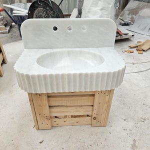 May include: A white marble sink with a scalloped edge and three faucet holes. The sink is mounted on a wooden pallet base. The sink has a rounded basin and a backsplash.