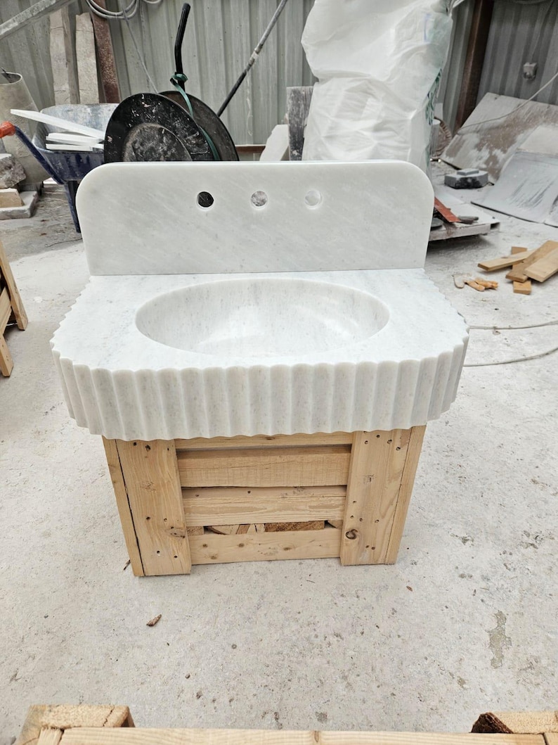 May include: A white marble sink with a scalloped edge and three faucet holes. The sink is mounted on a wooden pallet base. The sink has a rounded basin and a backsplash.