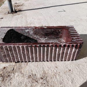 May include: A rectangular, carved stone sink with a dark red and gray marbled pattern. The sink has a central basin and a ridged exterior design. The sink is made of stone and is suitable for a bathroom.
