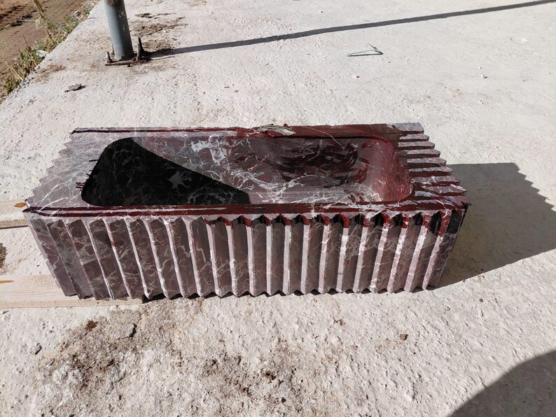 May include: A rectangular, carved stone sink with a dark red and gray marbled pattern. The sink has a central basin and a ridged exterior design. The sink is made of stone and is suitable for a bathroom.