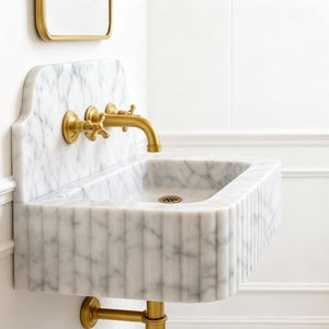 Puede incluir: Un lavabo de m&aacute;rmol montado en la pared con grifo y accesorios dorados. El lavabo tiene una cubeta rectangular con estr&iacute;as verticales en la parte delantera. El grifo tiene manijas en cruz. El lavabo est&aacute; sobre una pared blanca.