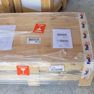 May include: A wooden crate wrapped in clear plastic, secured with FedEx tape. The crate has a "Fragile" sticker and several shipping labels. A stack of wooden bowls is visible in the background.