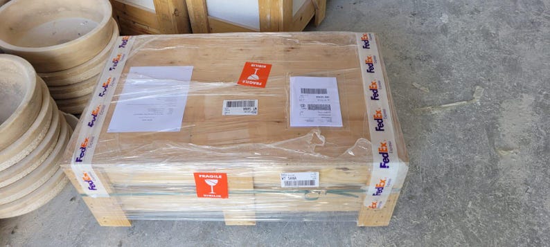 May include: A wooden crate wrapped in clear plastic, secured with FedEx tape. The crate has a "Fragile" sticker and several shipping labels. A stack of wooden bowls is visible in the background.