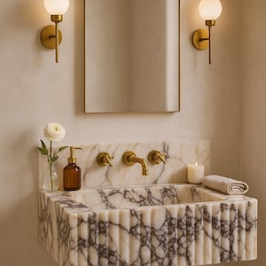 May include: A bathroom vanity with a marble sink, gold fixtures, and a matching arched mirror. Two sconces with globe lights flank the mirror. A small vase with a white flower and a candle are on the sink.