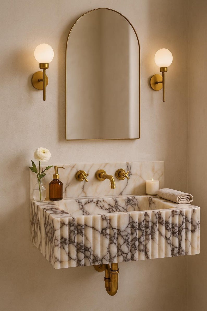 May include: A bathroom vanity with a marble sink, gold fixtures, and a matching arched mirror. Two sconces with globe lights flank the mirror. A small vase with a white flower and a candle are on the sink.