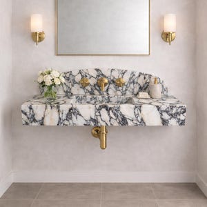 Könnte beinhalten: Ein luxuriöser Badezimmerwaschtisch mit Marmorplatte mit schwarzen Adern. Der Waschtisch hat goldene Armaturen, darunter Wasserhähne und ein Abflussrohr. Ein rechteckiger Spiegel hängt darüber, flankiert von zwei Wandleuchten. Eine Vase mit weißen Blumen.