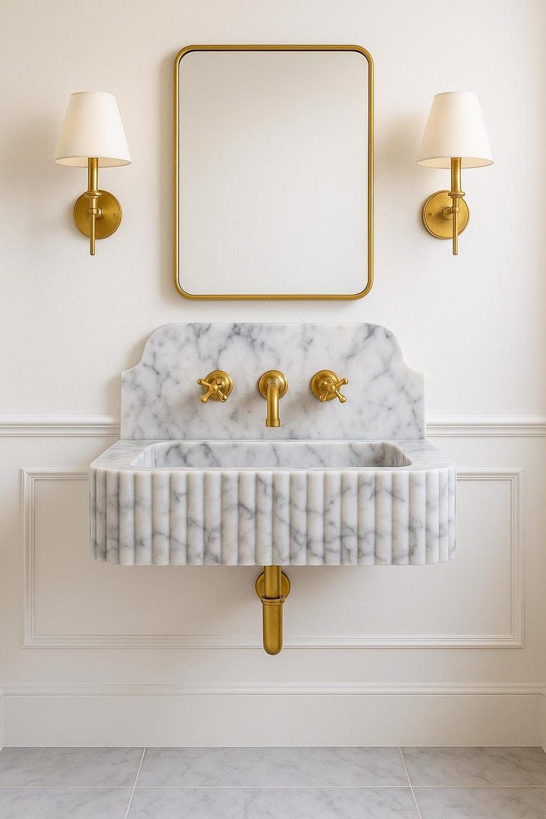 Puede incluir: Un lujoso conjunto de tocador de ba&ntilde;o con un lavabo de m&aacute;rmol, grifos dorados, un espejo rectangular y dos apliques de pared. El lavabo tiene un dise&ntilde;o acanalado y est&aacute; montado en una pared blanca.