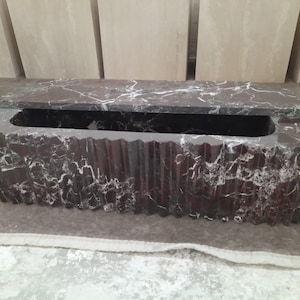 May include: A rectangular, dark red marble box with a lid. The box has a carved, textured exterior and a long, rectangular opening. White veining patterns the marble. The lid is partially open.