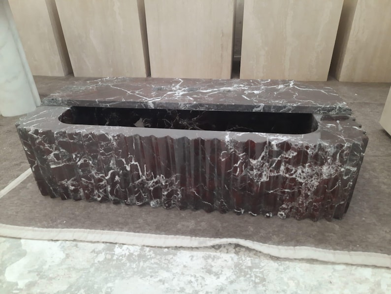 May include: A rectangular, dark red marble box with a lid. The box has a carved, textured exterior and a long, rectangular opening. White veining patterns the marble. The lid is partially open.