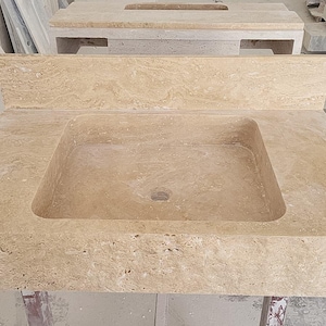Puede incluir: Un lavabo de ba&ntilde;o rectangular de piedra travertino beige que cuenta con una cubeta lisa y un salpicadero integrado a juego. La superficie de piedra natural muestra una textura porosa con sutiles variaciones de color y patr&oacute;n en la estructura.