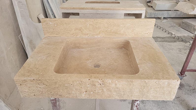 Puede incluir: Un lavabo de ba&ntilde;o rectangular de piedra travertino beige que cuenta con una cubeta lisa y un salpicadero integrado a juego. La superficie de piedra natural muestra una textura porosa con sutiles variaciones de color y patr&oacute;n en la estructura.
