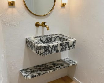 May include: A modern bathroom vanity set featuring a marble sink and matching shelf. The sink and shelf have a black and white veined pattern. A gold faucet, mirror, and sconces complete the design.